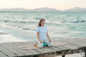 Somatisches Yoga: Jasmin sitzt auf einem türkisfarbenen Meditationskissen auf der ovalen FLOWALOHA-Matte am Meer